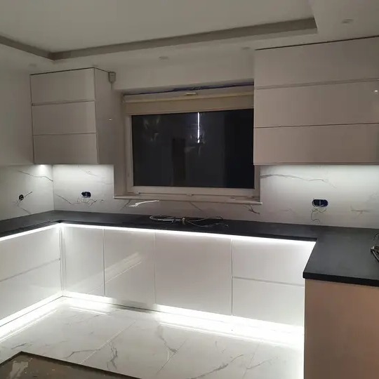 Nowoczesna, biała kuchnia z czarnym blatem i podświetleniem LED pod szafkami i nad blatem, marmurowe płytki na ścianie, widoczne gniazdka elektryczne.