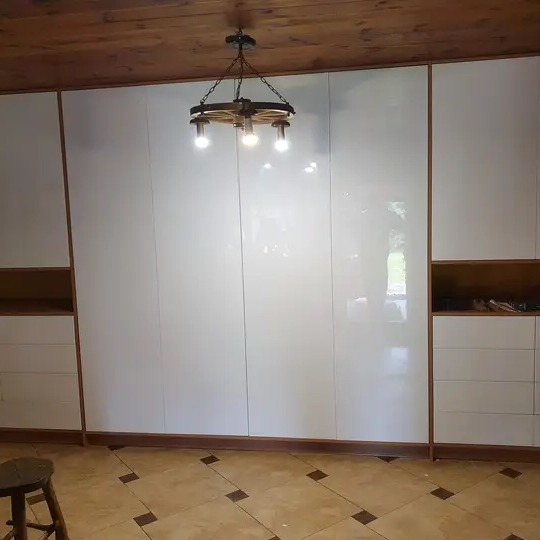 Biała, lakierowana szafa wnękowa z drewnianymi akcentami i szufladami, oświetlona dekoracyjną lampą z elementami koła w rustykalnym stylu, na tle drewnianego sufitu i wzorzystej podłogi.