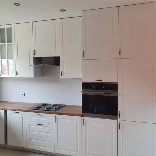 Biała kuchnia na wymiar z drewnianym blatem, czarnym piekarnikiem i szklanymi drzwiczkami szafek wiszących po lewej stronie.