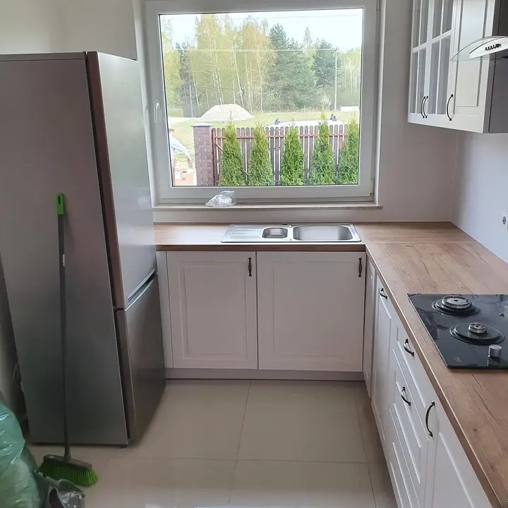 Jasna kuchnia z białymi szafkami i drewnianym blatem roboczym, widok na ogród przez okno, lodówka i mop w rogu, płytki podłogowe w odcieniach beżu.