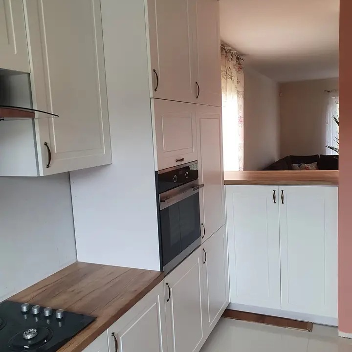 Jasna kuchnia z białymi szafkami z ciemnymi uchwytami i drewnianym blatem, z piekarnikiem w zabudowie i płytą grzewczą, widok na salon w tle.