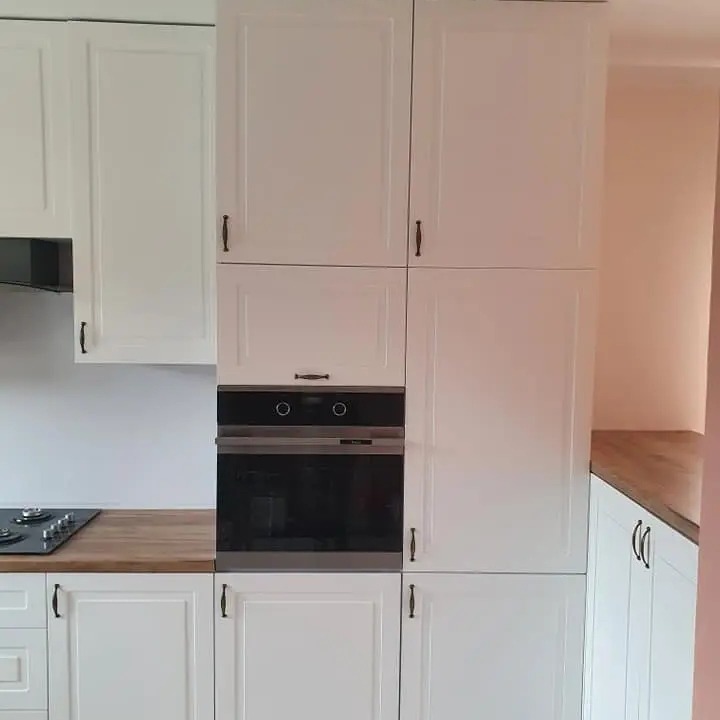 Biała zabudowa kuchenna z piekarnikiem w zabudowie, drewniany blat i płyta gazowa, proste uchwyty, minimalistyczny design.