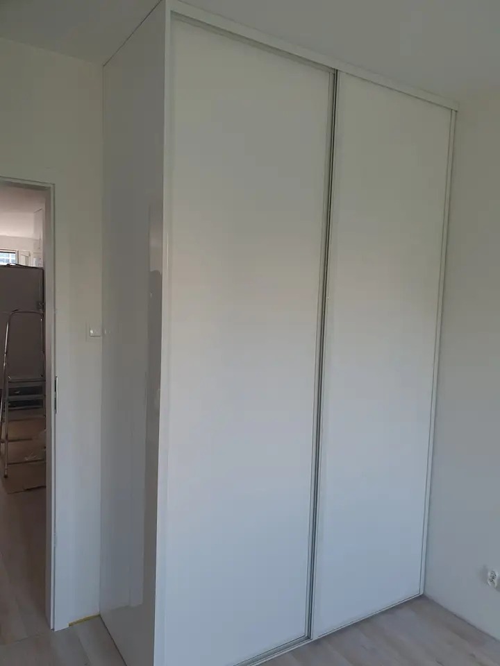 Biała szafa przesuwna wnękowa z dwoma drzwiami, widok z boku, w tle widoczne elementy wyposażenia wnętrza.
