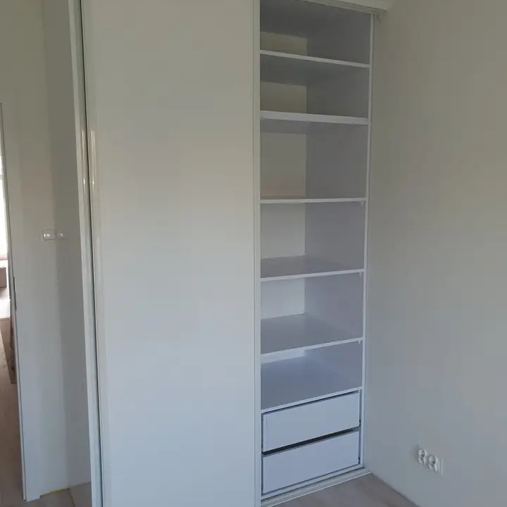 Biała szafa wnękowa z przesuwnymi drzwiami, widoczne półki i dwie szuflady, minimalistyczny design.