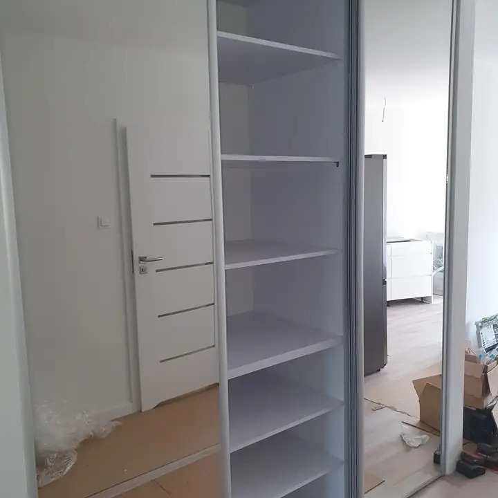 Biała szafa z lustrzanymi drzwiami przesuwnymi, widoczne puste półki i odbicie pokoju z drzwiami i kartonami.