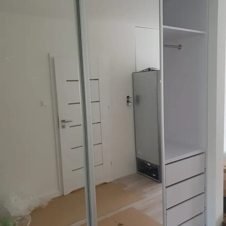 Biała szafa wnękowa z przesuwnymi drzwiami lustrzanymi, widoczne półki i szuflady, w tle drzwi i lodówka, podłoga zabezpieczona kartonem.