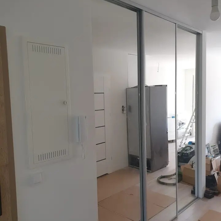 Szafa wnękowa z lustrzanymi drzwiami przesuwnymi w trakcie montażu, widoczne kartony i narzędzia, odbicie lodówki w lustrze.