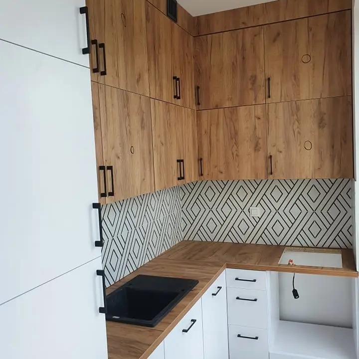 Nowoczesna kuchnia z drewnianymi szafkami górnymi, białymi szafkami dolnymi z czarnymi uchwytami i geometrycznym wzorem na ścianie nad blatem.