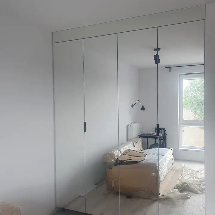 Biała szafa wnękowa z lustrzanymi drzwiami przesuwnymi, widoczne odbicie pokoju z meblami w folii ochronnej i kartonach, minimalistyczny design.