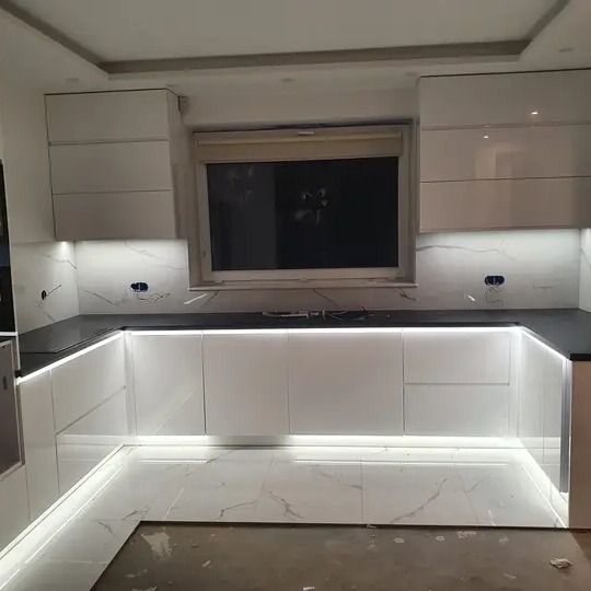 Biała, lakierowana kuchnia w kształcie litery U z podświetleniem LED pod szafkami i blatem, marmurowa podłoga, widoczne gniazdka elektryczne na ścianie.