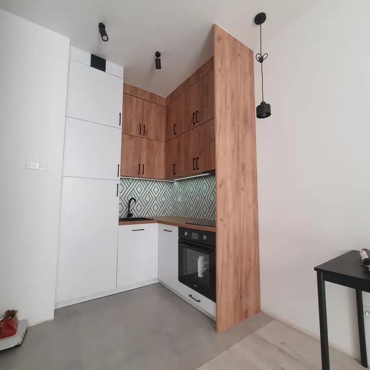 Nowoczesna kuchnia narożna z białymi frontami dolnymi i drewnopodobnymi górnymi szafkami, czarny zlew i bateria, geometryczne płytki nad blatem, oświetlenie punktowe i wisząca lampa.