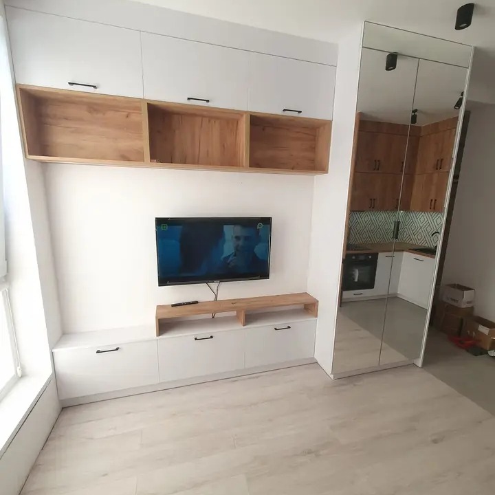 Białe szafki z drewnianymi akcentami nad telewizorem, odbicie kuchni w lustrzanych drzwiach szafy wnękowej.