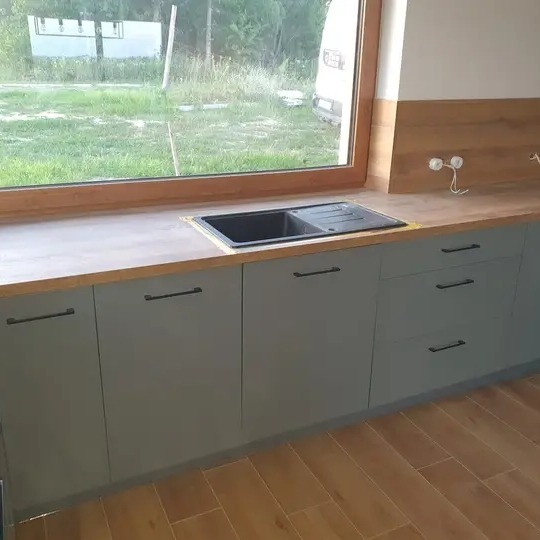Szare, minimalistyczne szafki kuchenne z czarnymi uchwytami i drewnianym blatem, widok na zewnątrz przez okno, Radomsko.