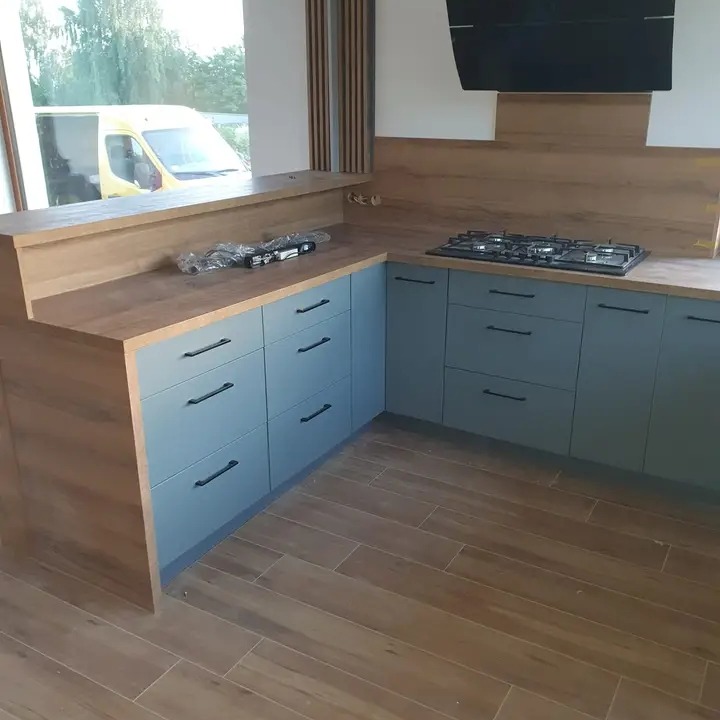 Narożna zabudowa kuchenna z szarymi frontami, drewnianym blatem i płytą gazową, widoczna z perspektywy podłogi w trakcie montażu, z narzędziami na blacie i transportowym autem za oknem.