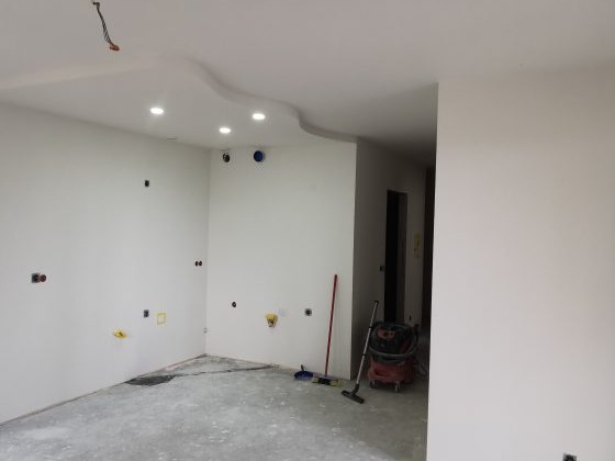 Wnętrze w trakcie prac wykończeniowych: ściany pomalowane na biało, widoczne punkty instalacji elektrycznej, listwa ozdobna na suficie, odkurzacz przemysłowy i mop stojące przy ścianie.