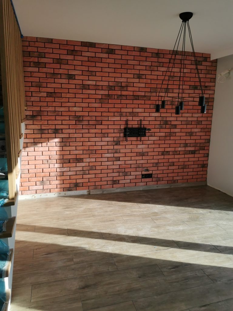 Ściana wykończona czerwoną cegłą klinkierową z czarnymi akcentami, widoczny uchwyt na telewizor i nowoczesna lampa wisząca z kilkoma czarnymi kloszami, podłoga wyłożona panelami imitującymi drewno.