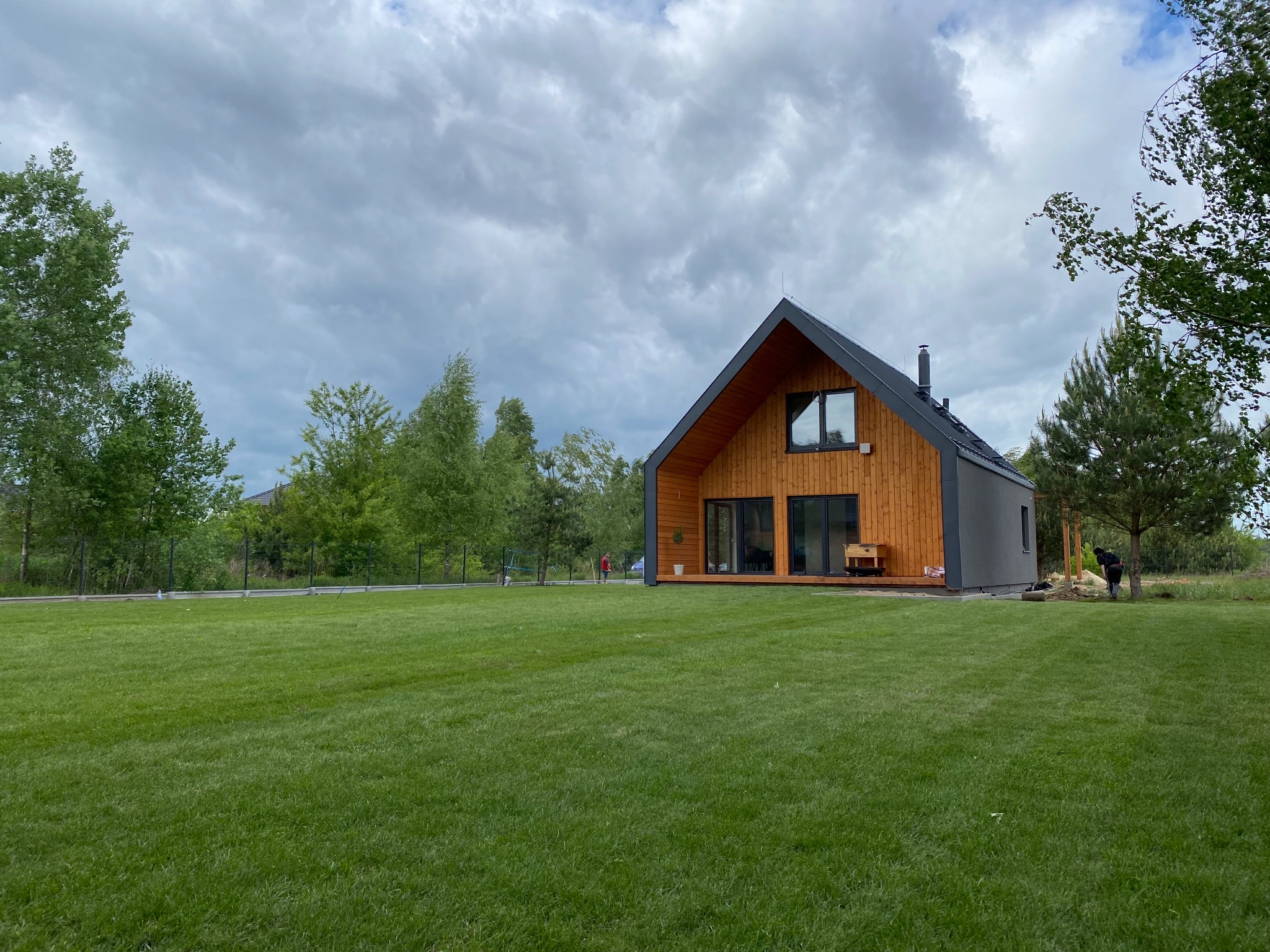 Nowoczesny dom z drewnianą elewacją i zadbanym trawnikiem. Osoba pracuje w ogrodzie, dodając naturalnego charakteru. Pochmurne niebo.