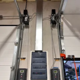 Szyna zasilająca i rozdzielnica elektryczna z automatyką do bramy garażowej, widoczne silniki z łańcuchami, sterowniki, przewody i elementy konstrukcyjne bramy.