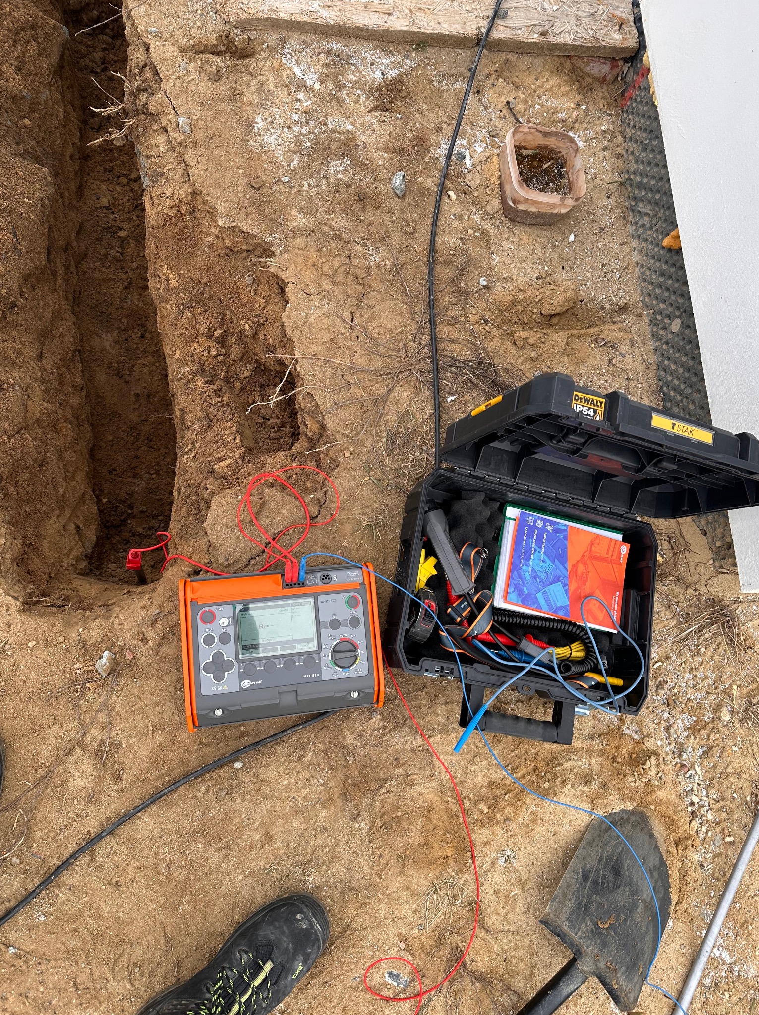 Pomarańczowy miernik elektryczny Comet MPI-520 z podłączonymi przewodami pomiarowymi, obok otwarta czarna skrzynka narzędziowa DeWalt TSTAK, szpadel i wykop w ziemi.