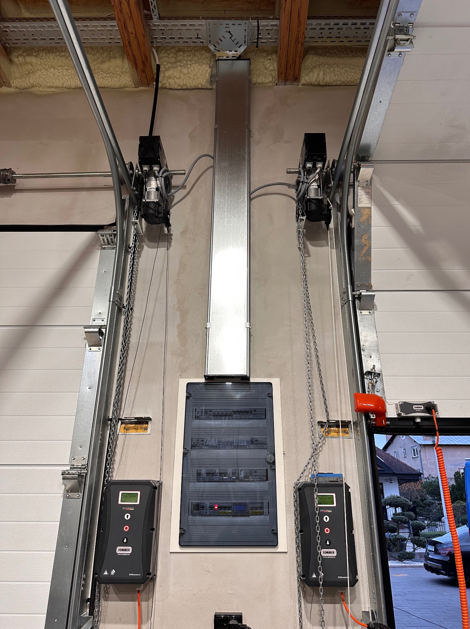 Szyna zasilająca i rozdzielnica elektryczna z automatyką do bramy garażowej, widoczne silniki z łańcuchami, sterowniki, przewody i elementy konstrukcyjne bramy.