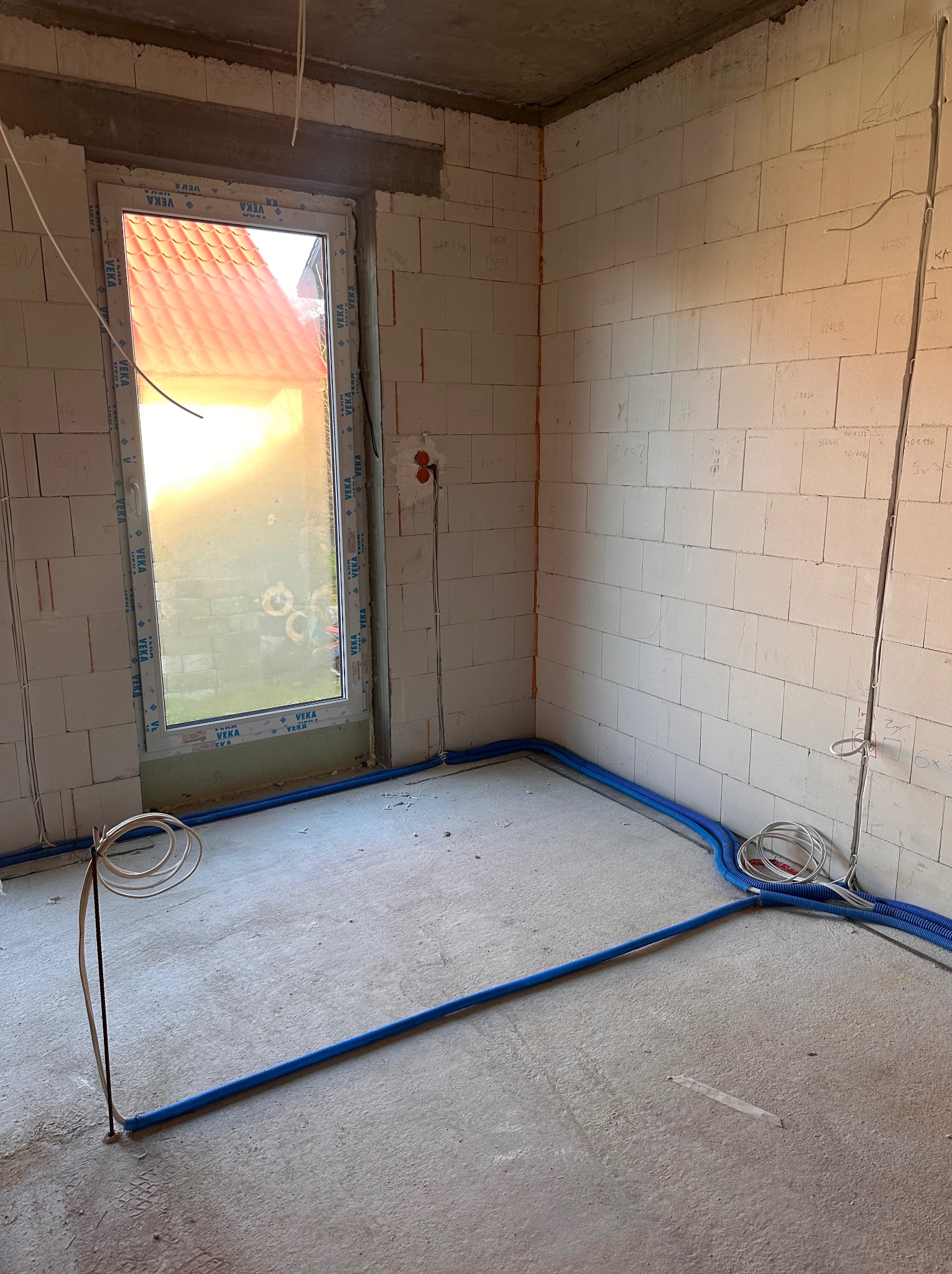 Surowy pokój z instalacją elektryczną w niebieskich rurach na podłodze, widoczne kable i puszki. Ściany z białego bloczka, okno z widokiem na dach.