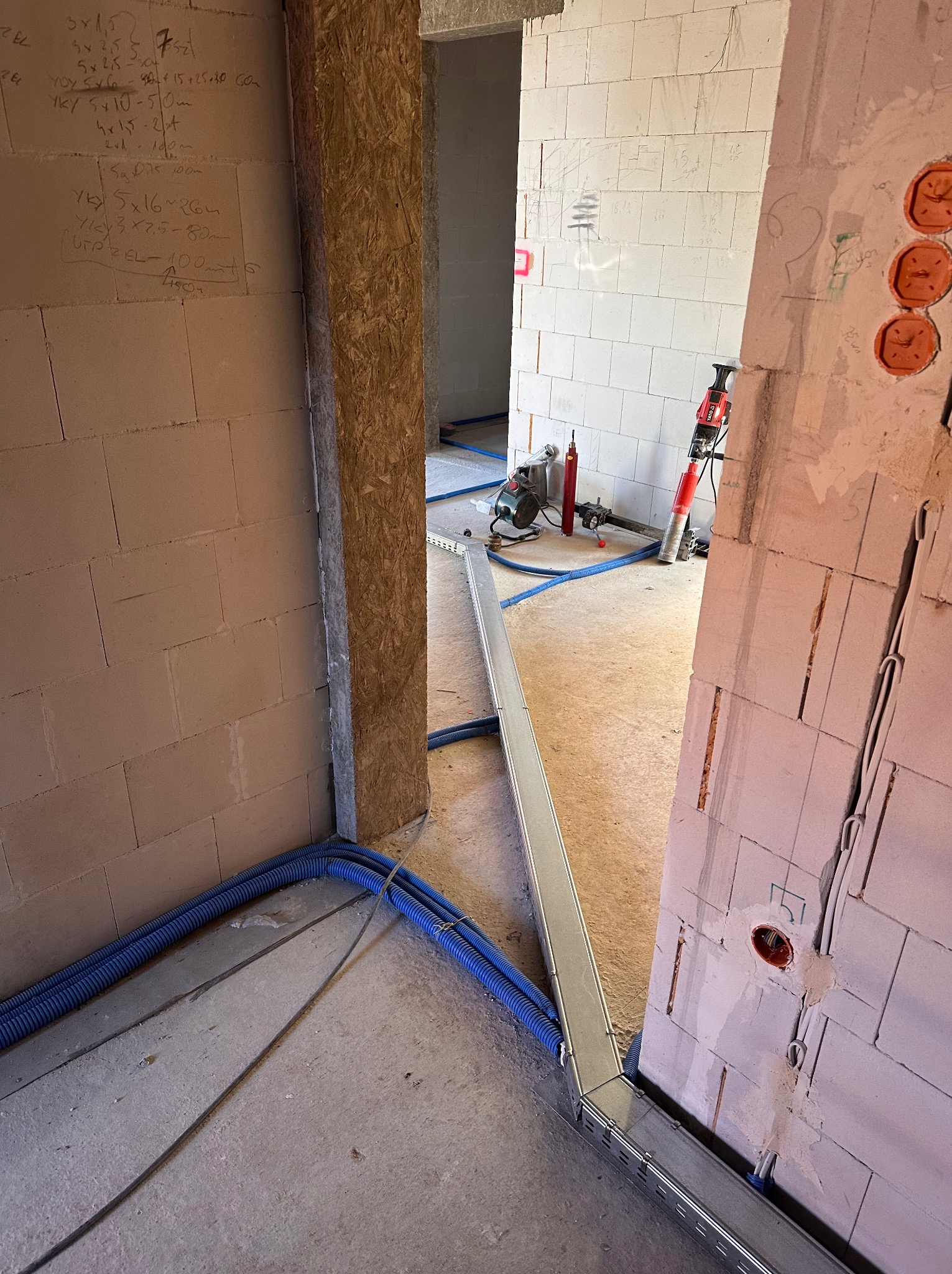 Instalacja elektryczna w trakcie budowy: niebieskie rury w korycie, puszki w ścianie z betonu komórkowego, narzędzia w tle. Widoczne notatki na ścianie.