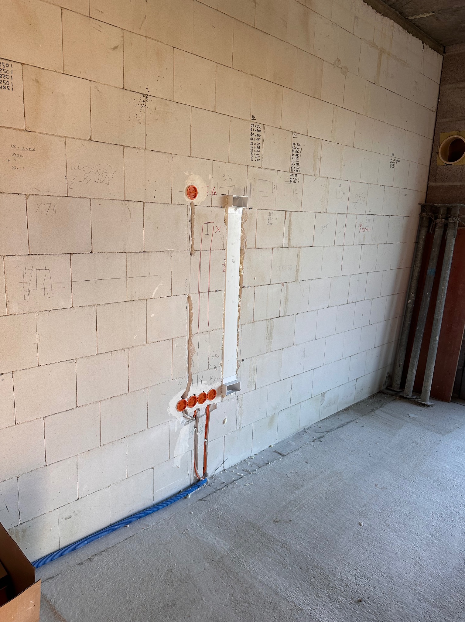 Ściana z betonu komórkowego z wyciętymi bruzdami pod kable i puszkami elektrycznymi. Widoczne przewody w kolorze pomarańczowym i niebieska rura karbowana.