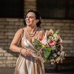 Karol Wierciński - Uśmiechnięta kobieta w złotej sukni z bukietem kwiatów i perłami na szyi, na tle ceglanej ściany.