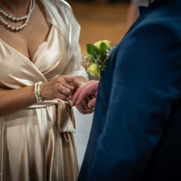 Karol Wierciński - Panna młoda w satynowej sukni i perłach zakłada obrączkę na palec pana młodego w granatowym garniturze, bukiecik żółtych róż.