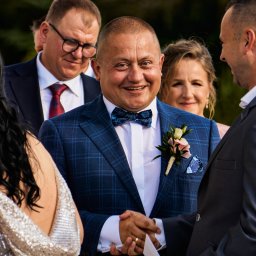 Karol Wierciński - Uśmiechnięty mężczyzna w niebieskim garniturze w kratę i z butonierką, witający się z gościem podczas uroczystości, w tle widoczni inni uczestnicy wydarzenia.