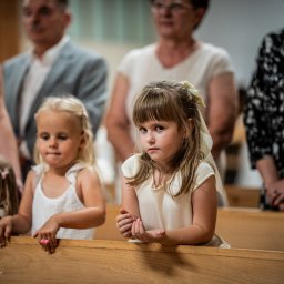 Karol Wierciński - Dziewczynka z kokardą we włosach, w białej sukience, siedzi w ławce kościelnej podczas uroczystości, zamyślona mina, na drugim planie rodzina i inne dzieci.