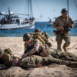 Karol Wierciński - Inscenizacja historyczna: żołnierze na plaży, jeden udziela pomocy rannemu, w tle łodzie na morzu.