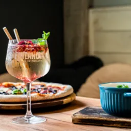 Drink Finlandia z owocami i miętą w kieliszku, pizza na drewnianej desce i niebieska miseczka na drewnianej podkładce, na drewnianym stole.