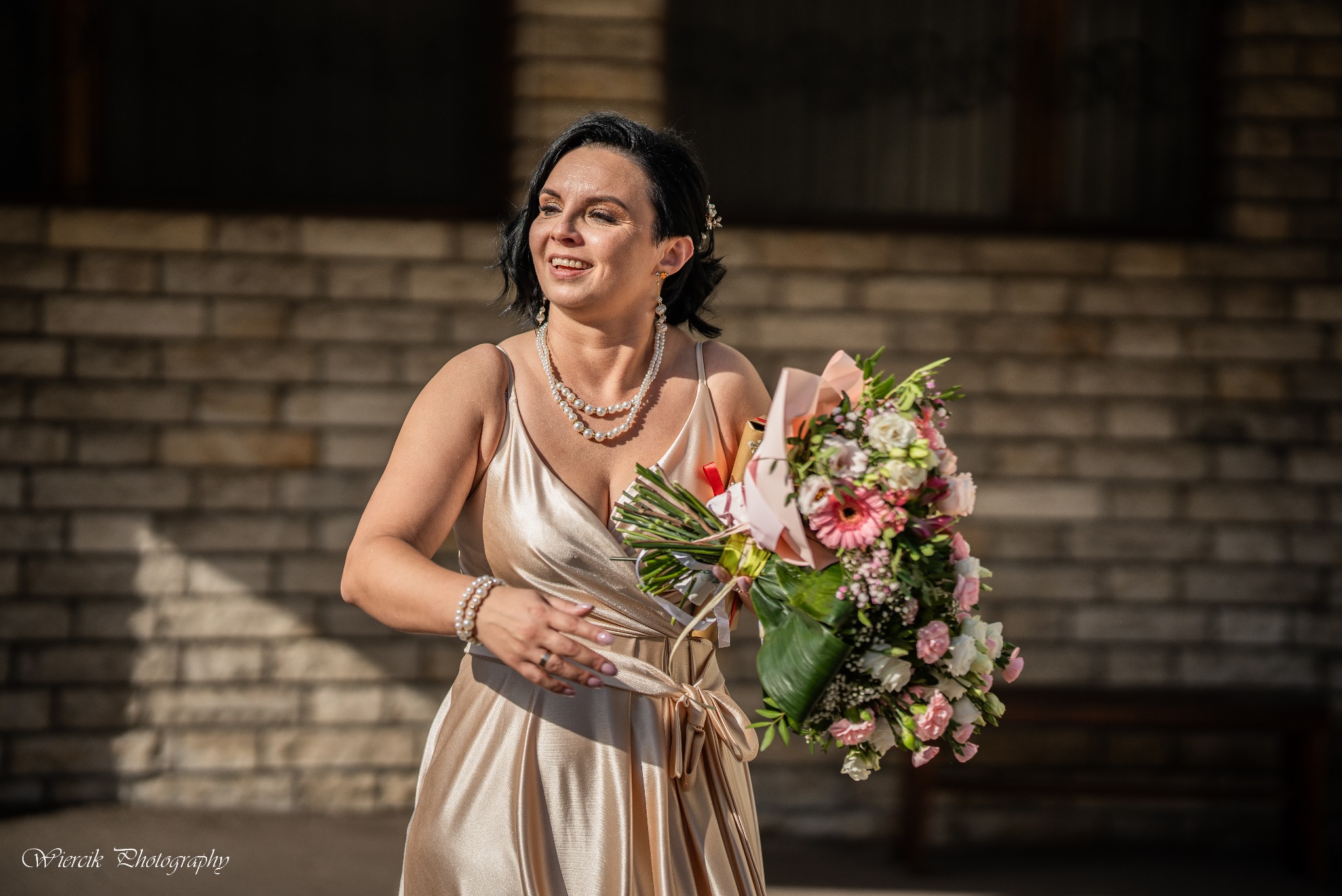 Uśmiechnięta kobieta w złotej sukni z bukietem kwiatów i perłami na szyi, na tle ceglanej ściany.