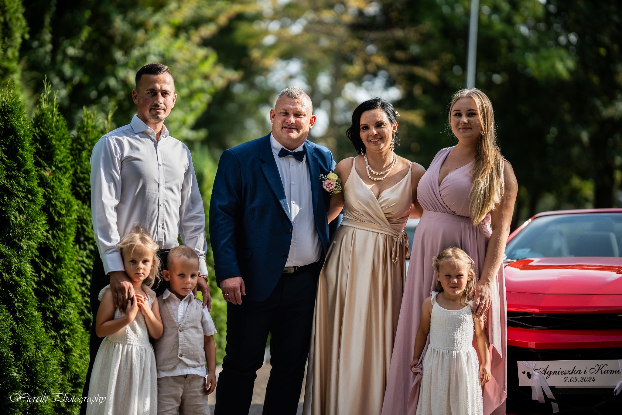 Grupa osób w strojach wizytowych, w tym para młoda z dziećmi i gośćmi, pozuje do zdjęcia na tle zieleni i czerwonego samochodu z dekoracją ślubną z datą 7.09.2024.