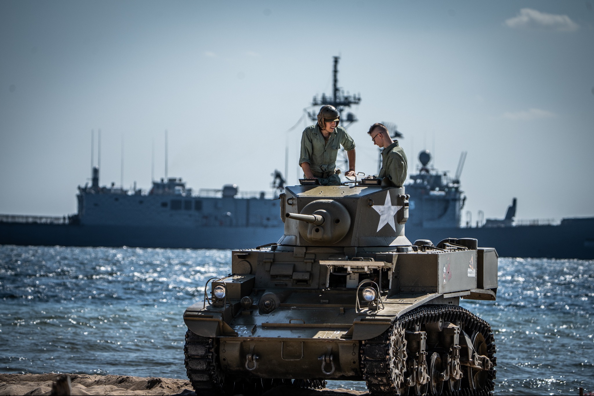Dwóch mężczyzn w mundurach na czołgu M3 Stuart z gwiazdą, na tle morza i okrętu wojennego podczas rekonstrukcji historycznej.