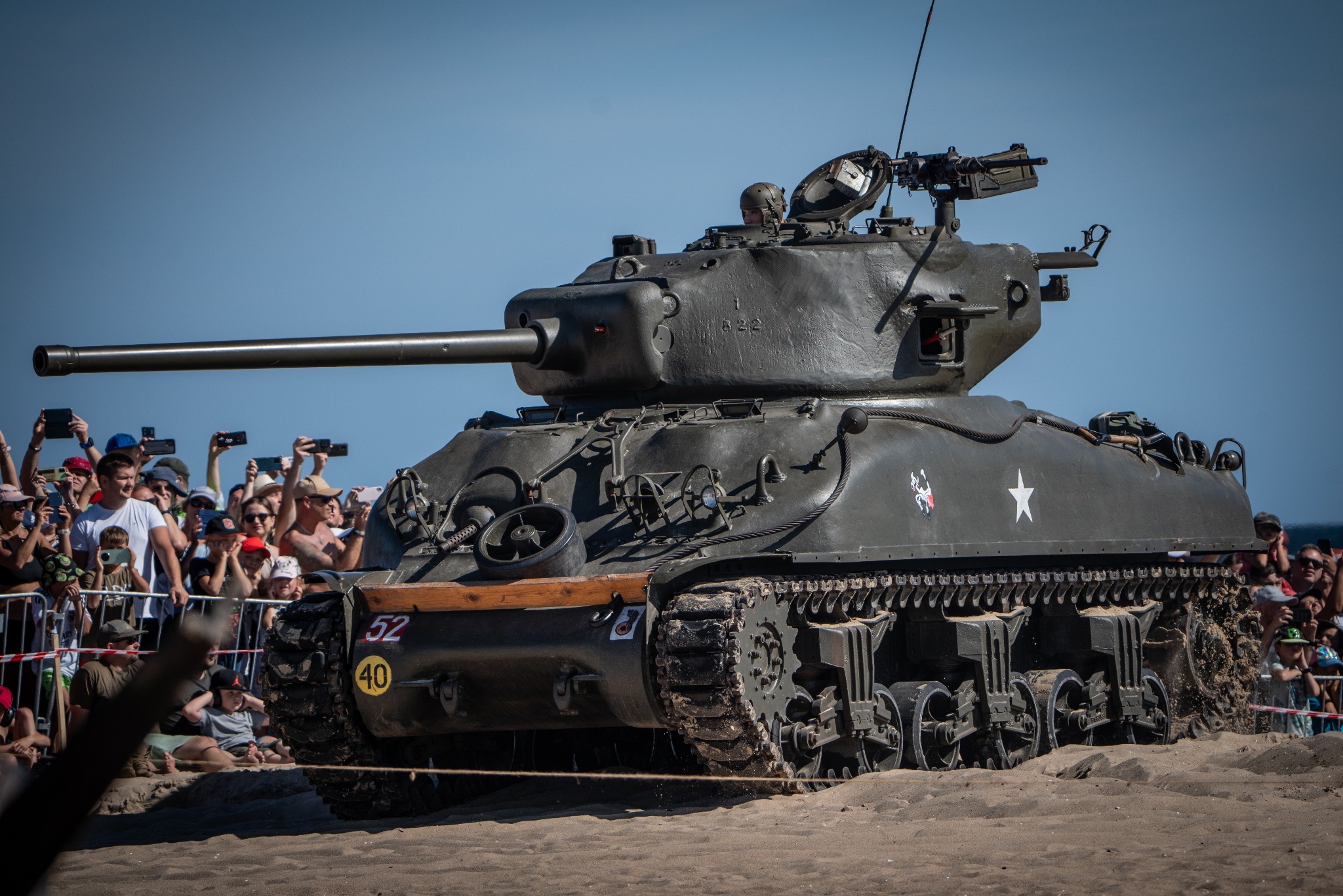 Czołg Sherman M4A3 na plaży, otoczony przez tłum ludzi z telefonami; widoczny numer 52 i biała gwiazda na pancerzu.