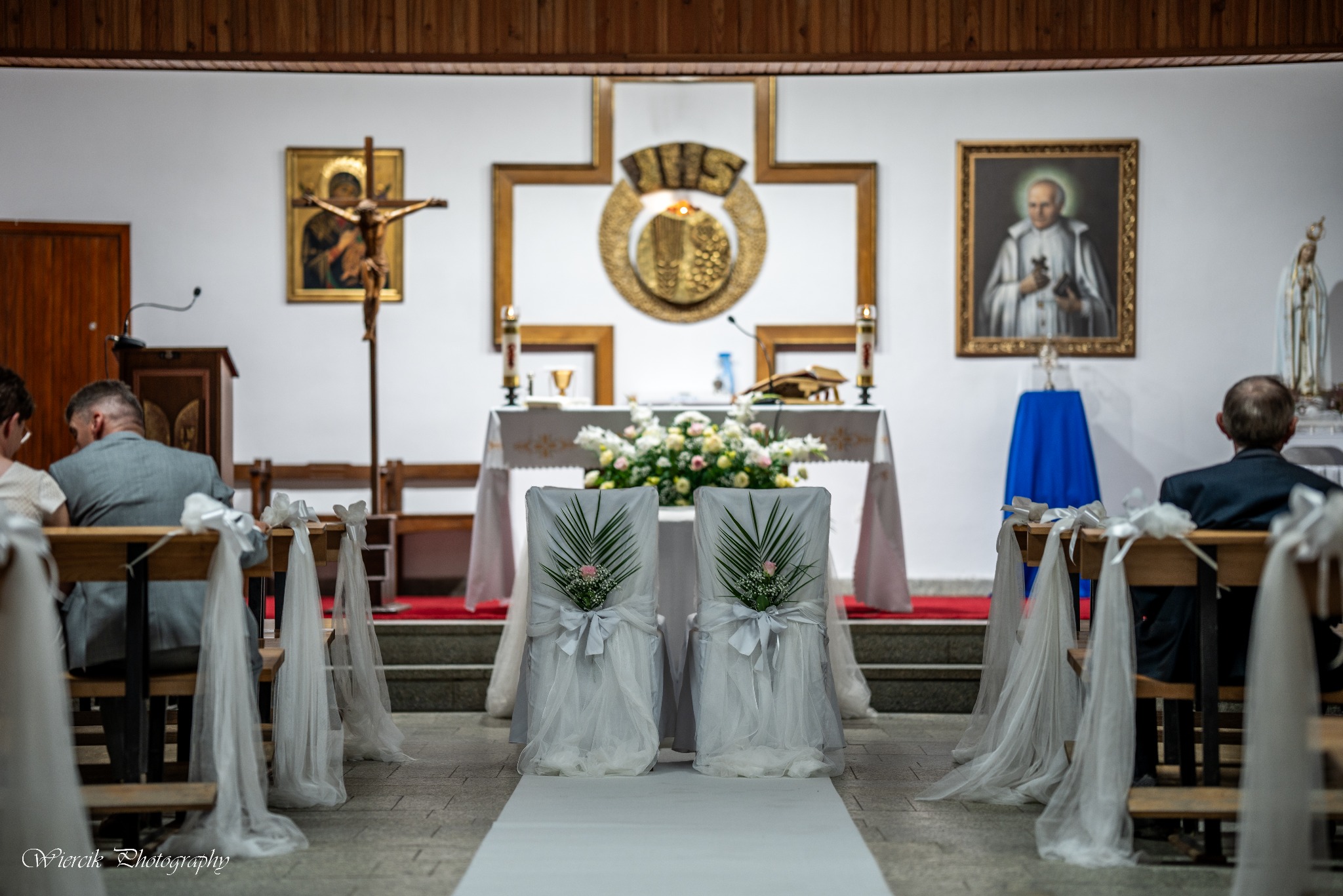 Udekorowane krzesła dla pary młodej w kościele, biały dywan, kwiaty na ołtarzu, wierni siedzący w ławkach, obrazy religijne na ścianach.