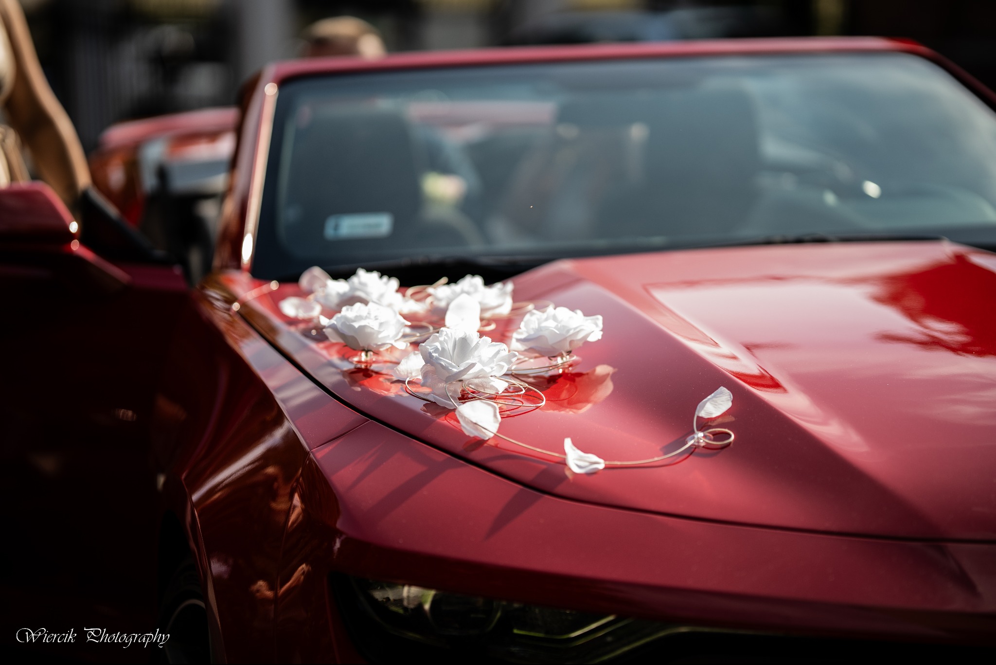 Czerwony samochód udekorowany białymi kwiatami i złotymi obrączkami na masce, widoczny fragment otwartych drzwi i wnętrza pojazdu.