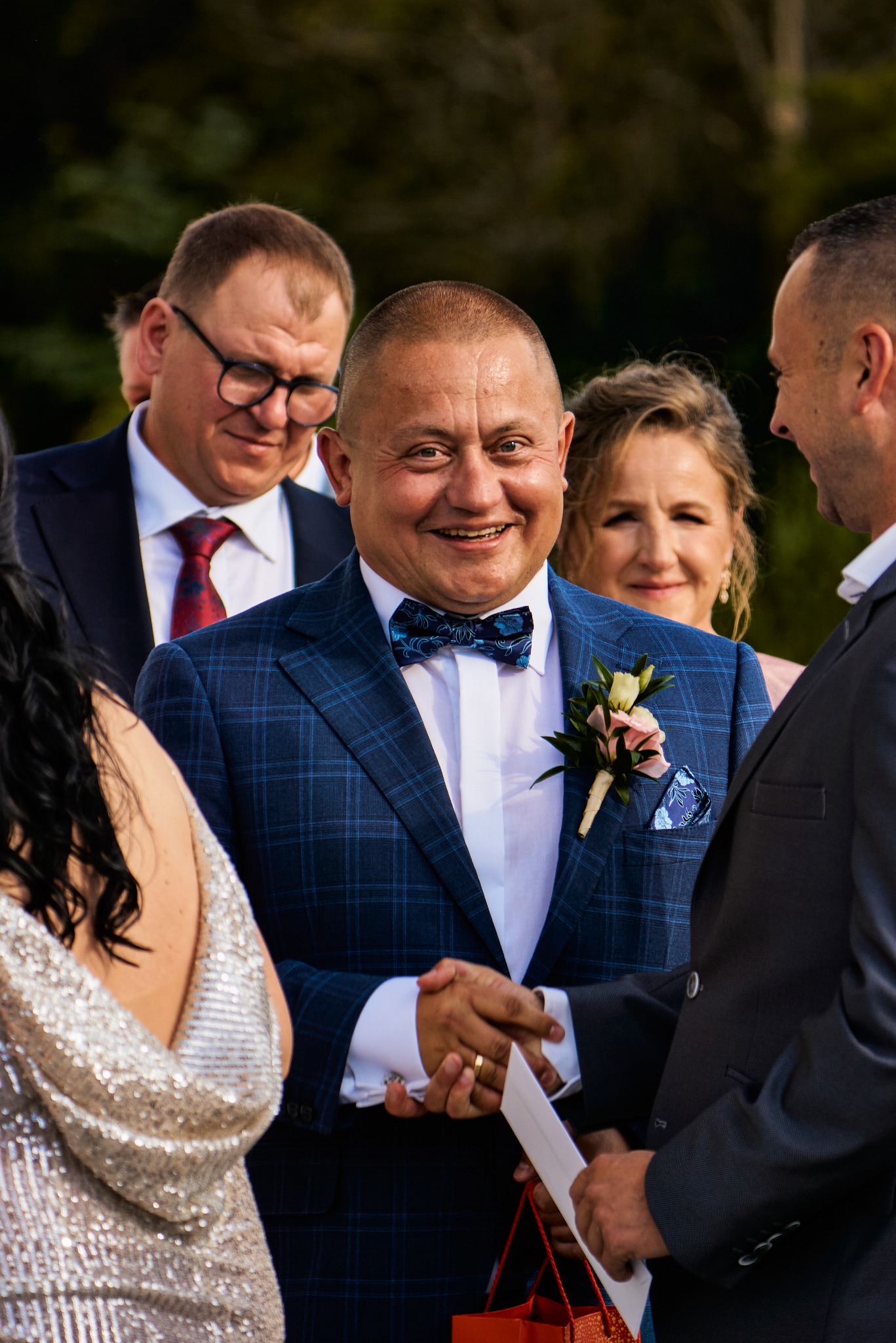 Uśmiechnięty mężczyzna w niebieskim garniturze w kratę i z butonierką, witający się z gościem podczas uroczystości, w tle widoczni inni uczestnicy wydarzenia.