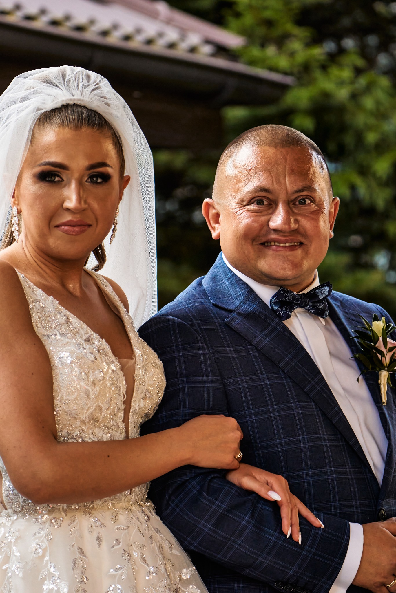 Para młoda: uśmiechnięta panna młoda w bogato zdobionej sukni i welonie, oraz pan młody w granatowym garniturze w kratę z muszką, pozują do portretu w plenerze.