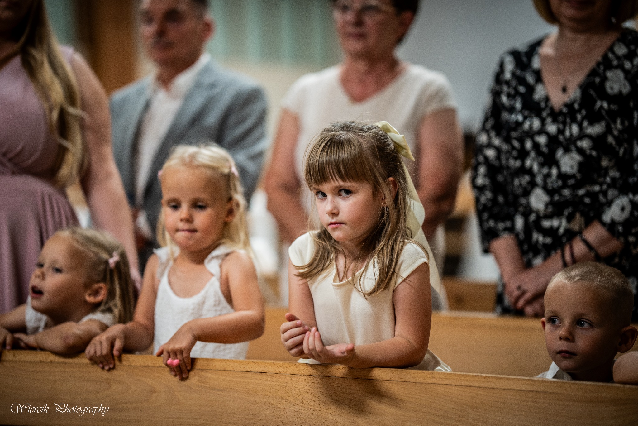 Dziewczynka z kokardą we włosach, w białej sukience, siedzi w ławce kościelnej podczas uroczystości, zamyślona mina, na drugim planie rodzina i inne dzieci.