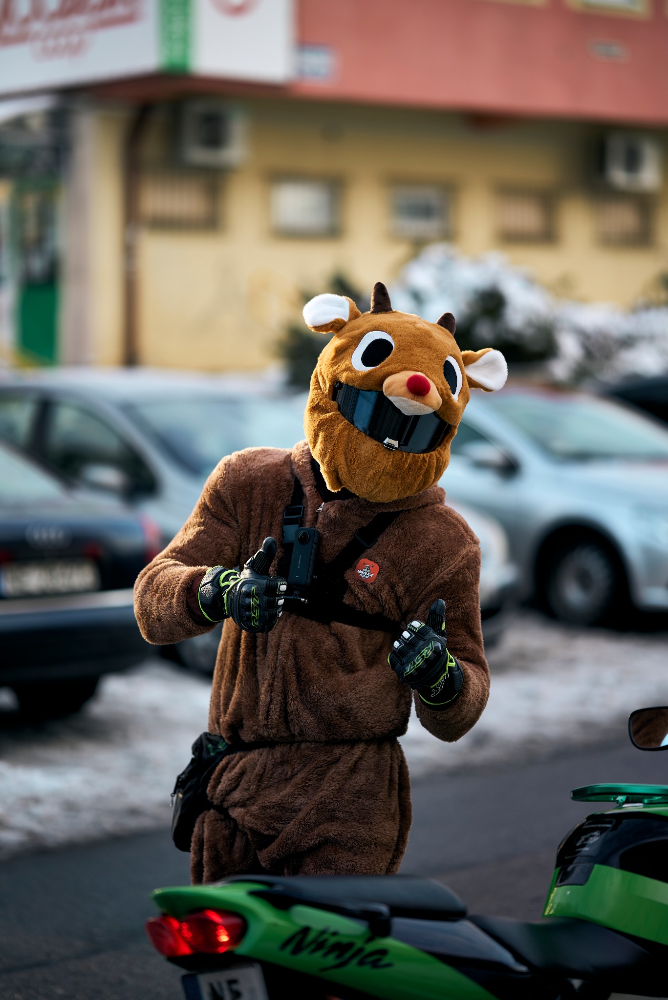 Osoba w kostiumie renifera z kaskiem, dająca dwa kciuki w górę obok zielonego motocykla Ninja, na tle miejskiego krajobrazu z samochodami.