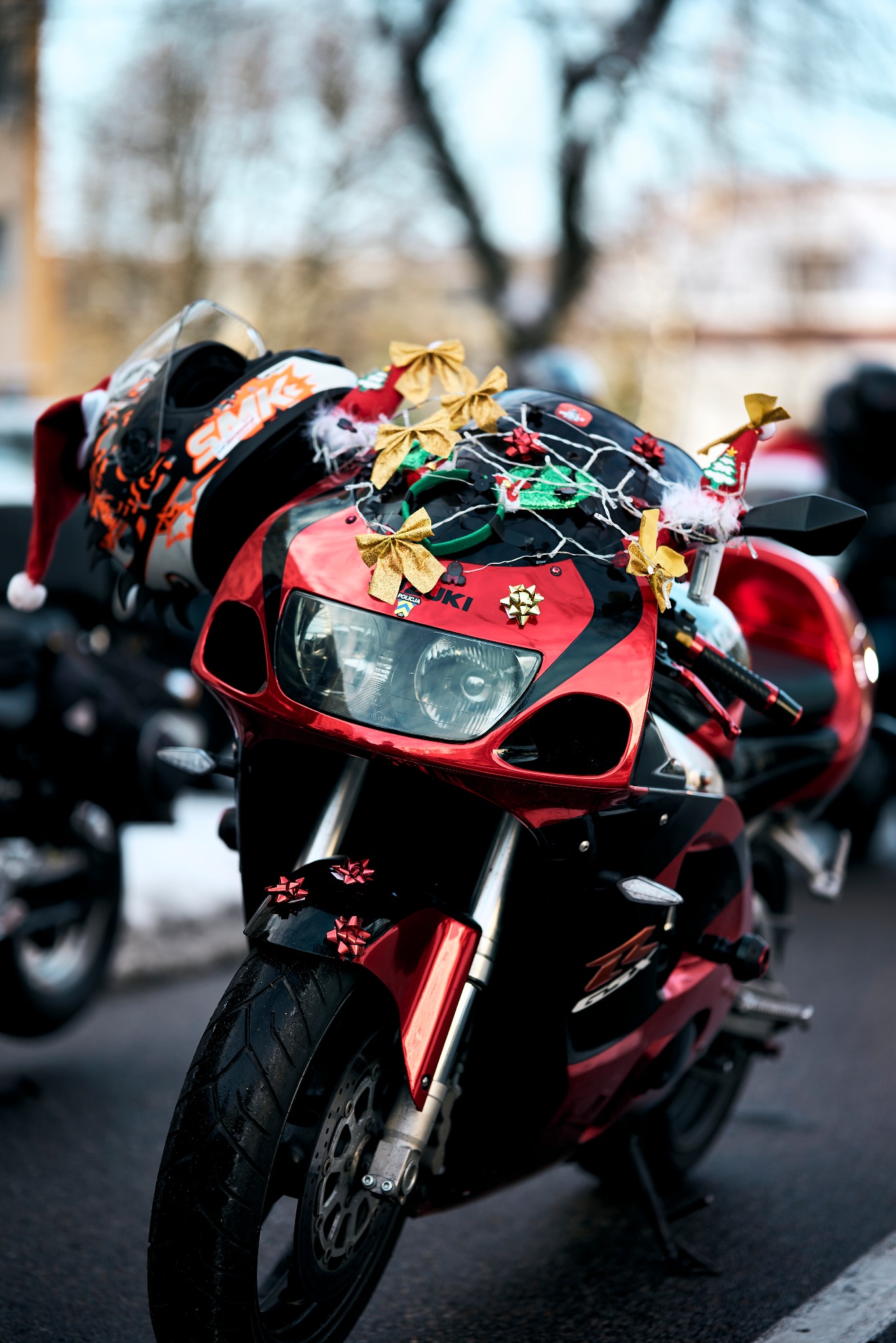 Czerwony motocykl Suzuki udekorowany świątecznymi ozdobami, w tym złotymi kokardkami, lampkami i czapką Świętego Mikołaja na kasku.