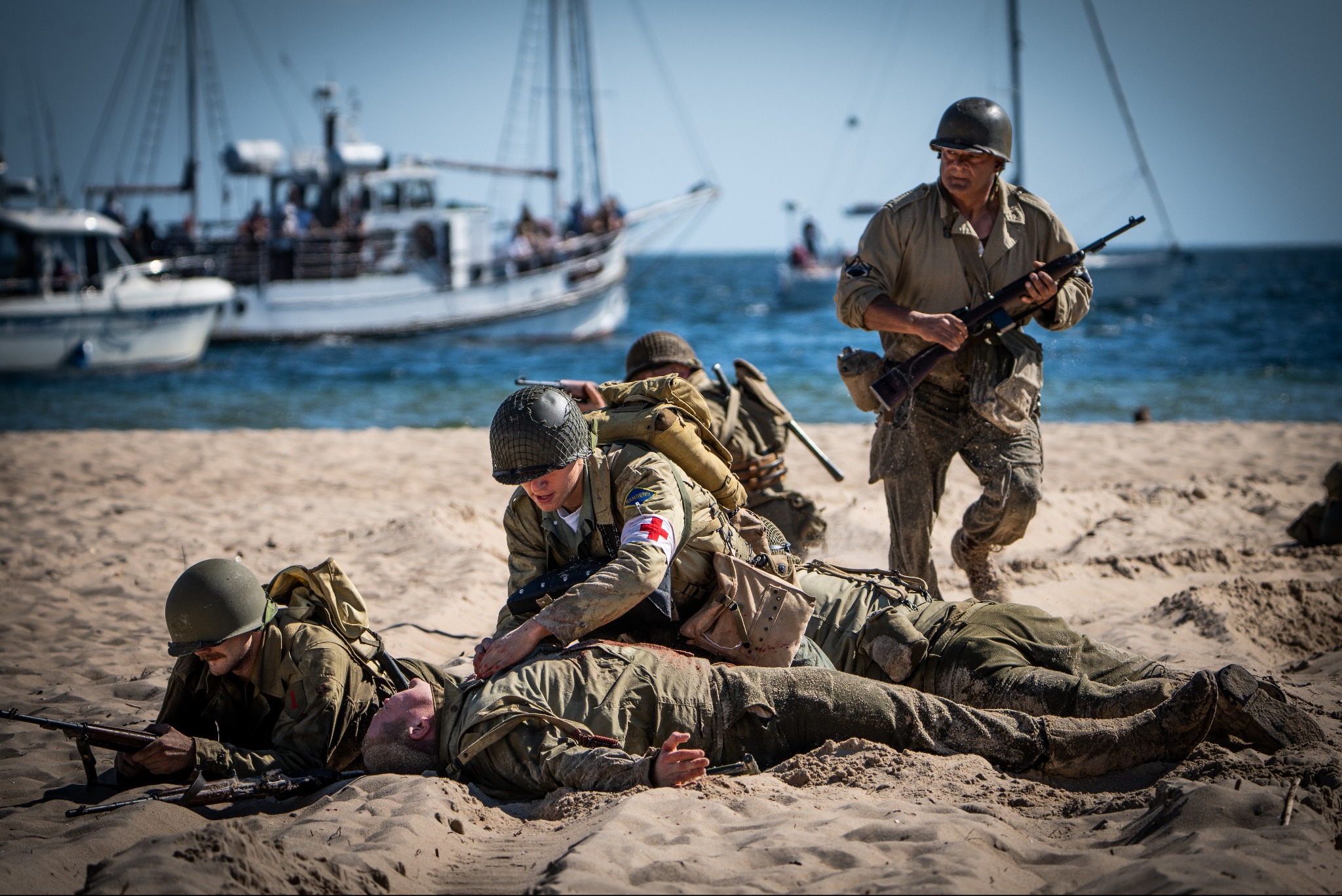 Inscenizacja historyczna: żołnierze na plaży, jeden udziela pomocy rannemu, w tle łodzie na morzu.