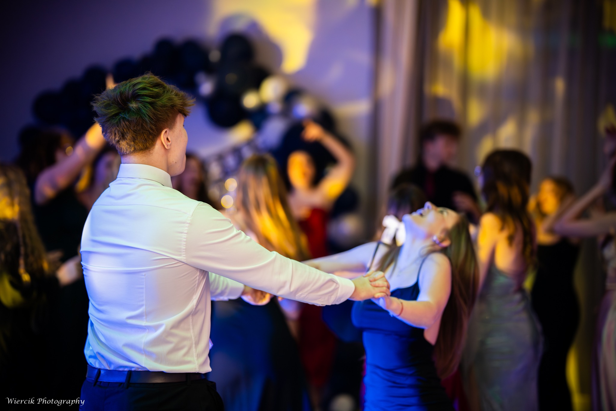 Para tańczy na parkiecie podczas uroczystości. W tle widoczni inni goście i dekoracje balonowe. Mężczyzna w białej koszuli, kobieta w niebieskiej sukience.