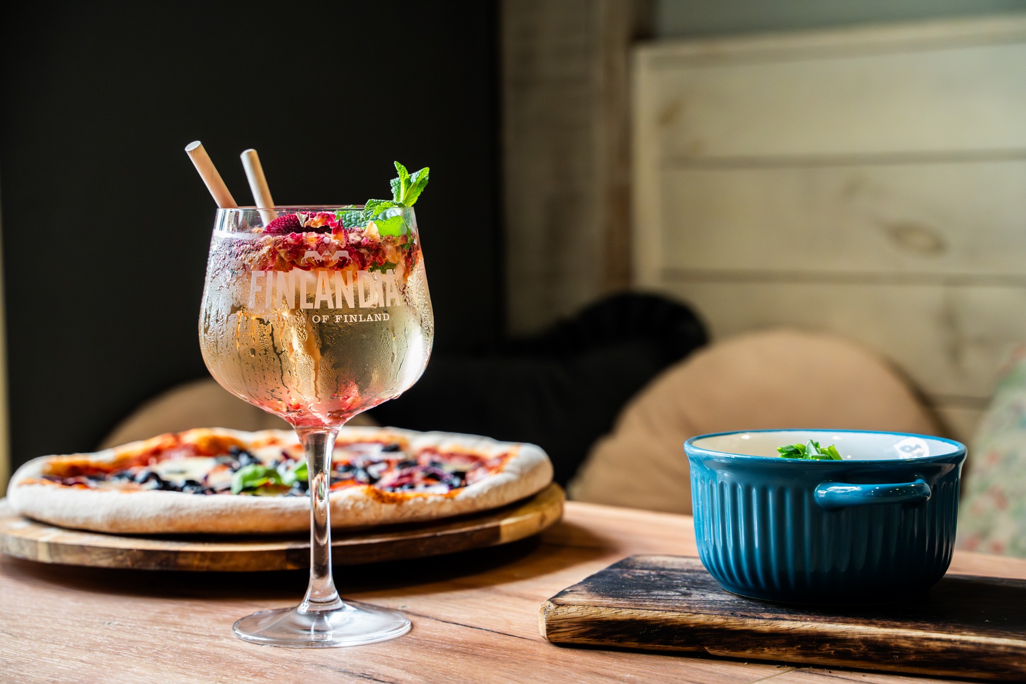 Drink Finlandia z owocami i miętą w kieliszku, pizza na drewnianej desce i niebieska miseczka na drewnianej podkładce, na drewnianym stole.