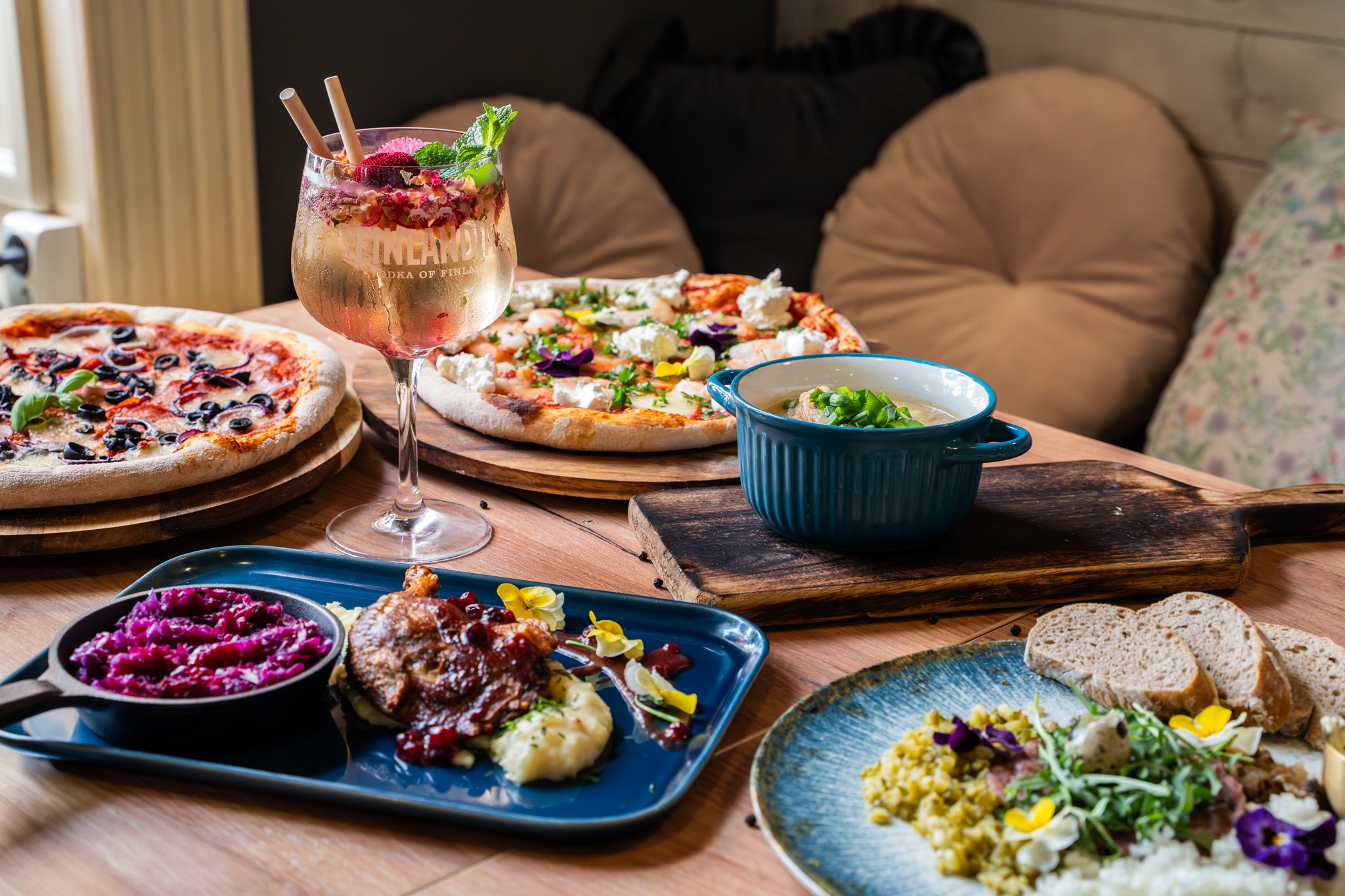 Bogato zastawiony stół z pizzą, zupą, koktajlem i daniami mięsnymi w Elblągu. Różnorodne potrawy na drewnianym stole w restauracyjnym wnętrzu.