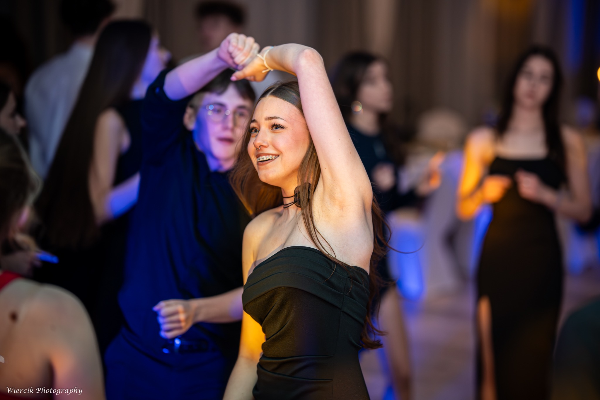 Uśmiechnięta brunetka w czarnej sukience tańczy na imprezie, widoczne aparaty ortodontyczne i piercing w nosie, w tle rozmazane sylwetki innych osób.