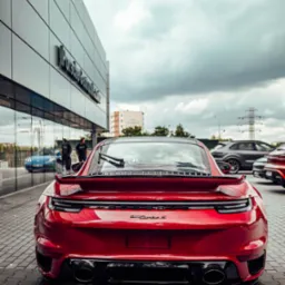 Czerwone Porsche 911 Turbo S zaparkowane przed salonem Porsche Centrum, widok z tyłu, zarys sylwetek w odbiciu szyby.